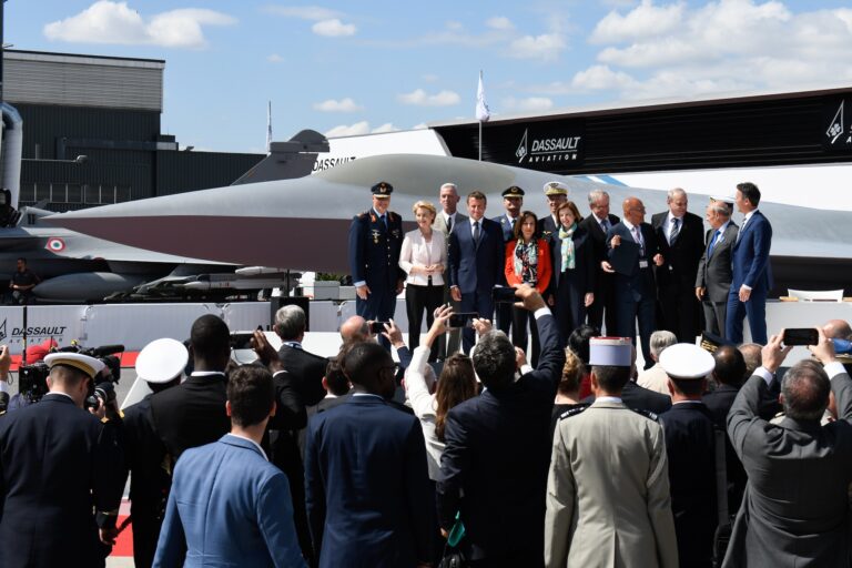 French and German officials stand on a podium in front of a grey fighter jet. They face a crowd of people, some hold up phones to take photos. It is a sunny day.