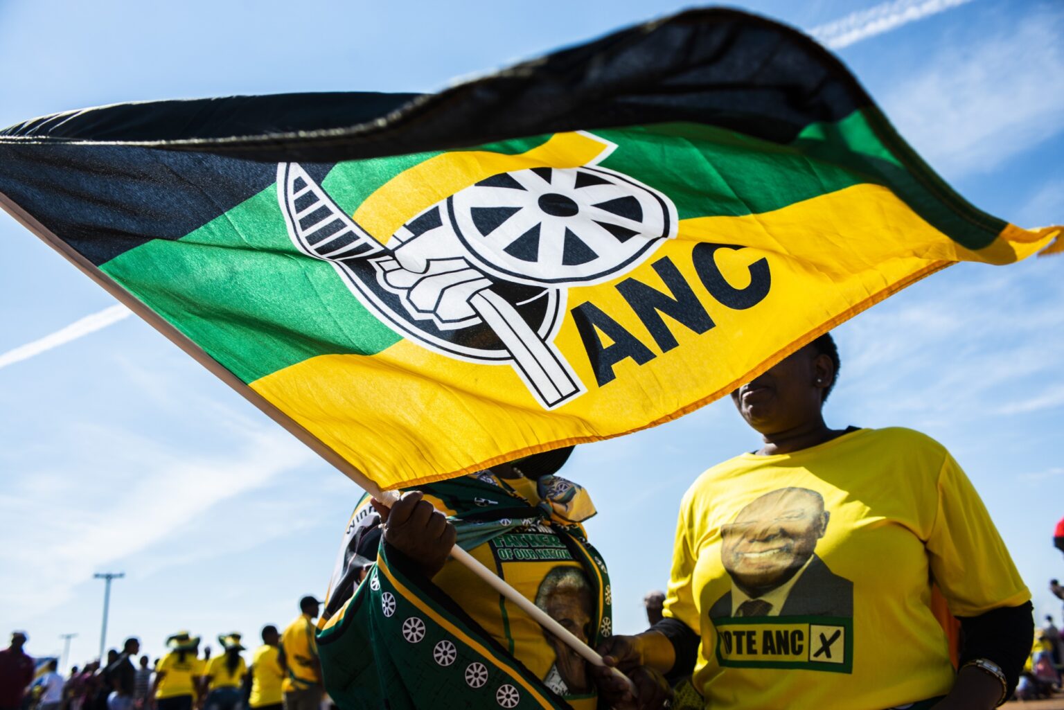 Two ANC supporters wearing African National Congress party T-shirts wave a large flag, obscuring their faces. One has a ANC flag draped over their shoulders. In the background more ANC supporters wearing party colours stand at a distance.