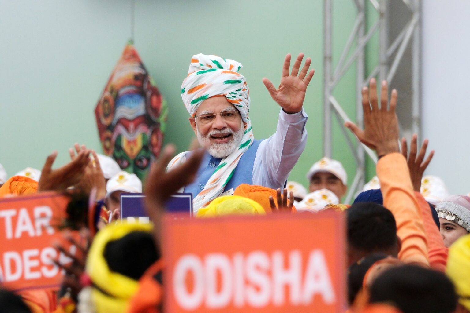 India's PM Narendra Modi celebrating India's Independence Day at a rally in 2022. He wears a white turban with a green and orange pattern and is waving to the crowd. He stands in front of a mint green wall. The crowd wave back and hold orange placards, they wear orange and yellow.