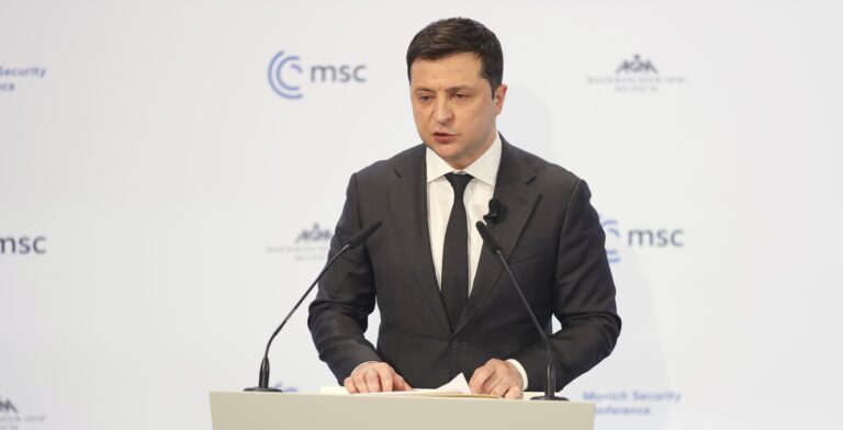 Ukrainian President Volodymyr Zelensky delivers a statement during the Munich Security Conference in Munich, Germany. Zelensky wears a dark suit and tie and stands behind a white podium with the logo of the Munich Security Conference.