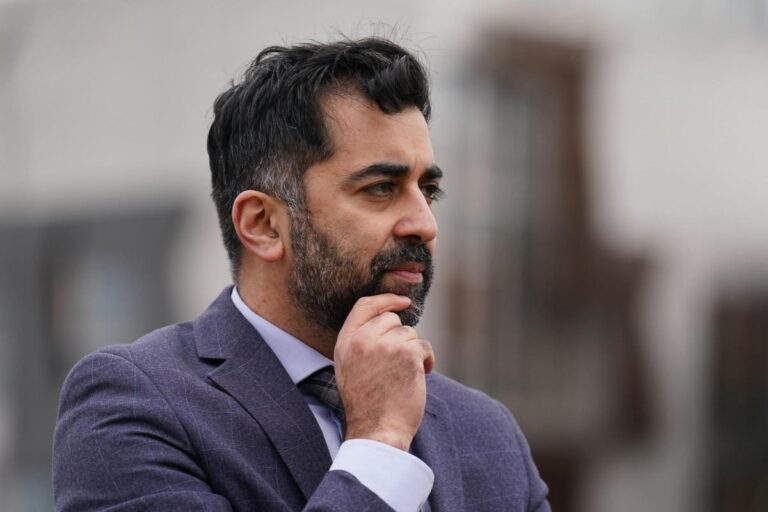 Humza Yousef turned slightly to his left looking into the distance, his right hand rests on his chin. He wears a checked blue-grey suit and looks thoughtful. The background is grey and out of focus, and gives the impression of an outdoor urban background.