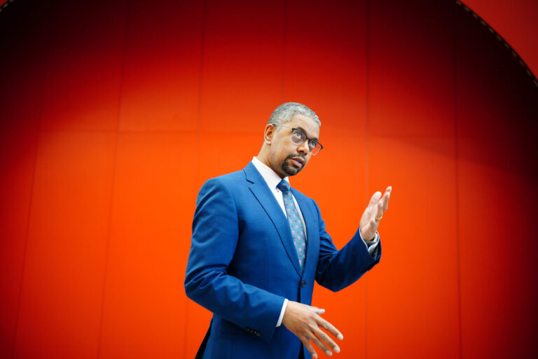Vaughan Gething poses for a photo, in a lecture hall at Cardiff University, after being elected as the next Welsh Labour leader and First Minister of Wales, in Cardiff. He wears a blue suit and light blue tie and had his hands clasped in front of him. He stands in front of a bright red background, which fades to black at the edges of the picture.