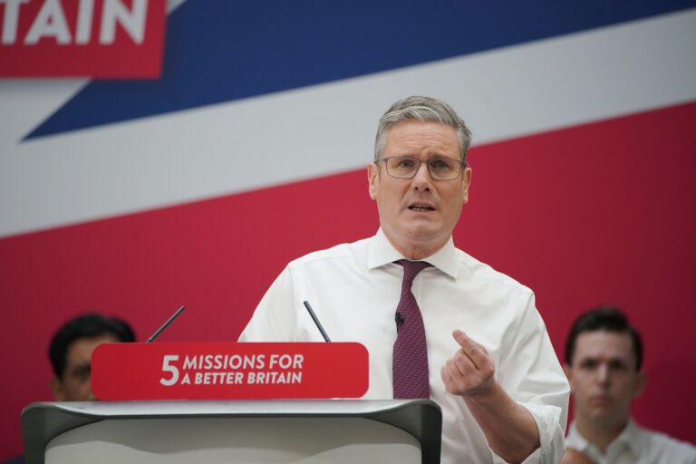 Leader of the Labour Party, Sir Keir Starmer, delivers a speech in a white shirt and red tie. He stands behind and slightly to one side of a podium which is topped with a sign that reads "5 missions for a better Britain" in white letters on a red background. The background shows part of a Union Jack flag and two male audience members watching from behind Keir.